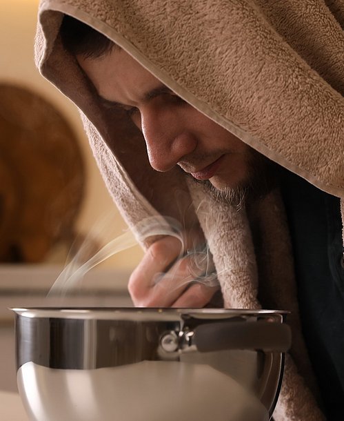 Hausmittel können bei der Behandlung von Bronchitis unterstützen. Mann mit Bronchitis inhaliert Kochsalzlösung am Küchentisch.