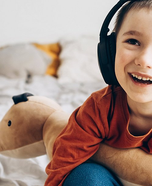 Wenn Kinder eine Erkältung haben, können Tipps gegen Langeweile, die Zeit angenehmer gestalten. Kind sitzt auf einem Bett und hat Kopfhörer auf.