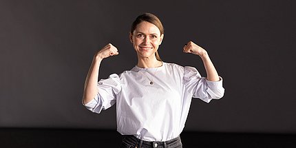 Frau mit angewinkelten Armen zur Demonstration von Stärke vor einem dunklen Hintergrund