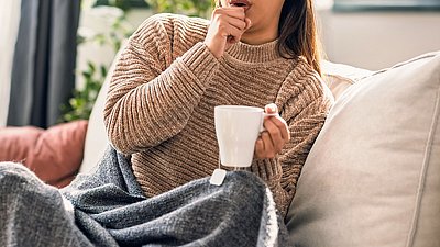 Frau sitzt mit einem Tee auf der Couch und hustet.